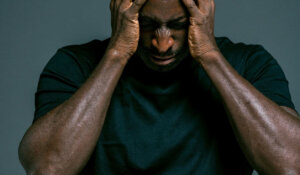 A bald man in a black shirt standing against a dark background, holding his head in distress, conveying emotional overwhelm and anger management struggles.